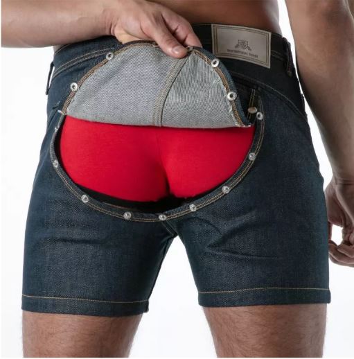 A close-up of a person wearing TOF Paris Raw Denim Backless Shorts Blue with a large rear opening, shown pulling the back of the shorts open to reveal bright red underwear underneath. A hand is visible pulling the fabric, and the person's legs are shown against a plain white background.